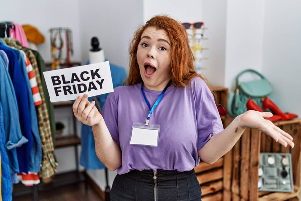 Молода руда жінка тримає чорний п'ятничний банер в роздрібному магазині, святкуючи досягнення з щасливою посмішкою і виразом переможця з піднятою рукою
  - Фото, зображення