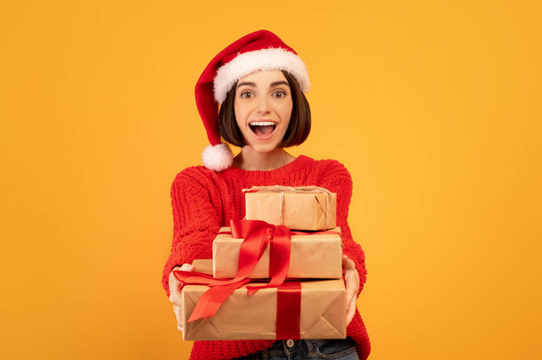 Emocionální šťastná žena v Santa klobouku drží dárkové krabice a roztahuje je na kameru přes žluté pozadí studia - Fotografie, Obrázek