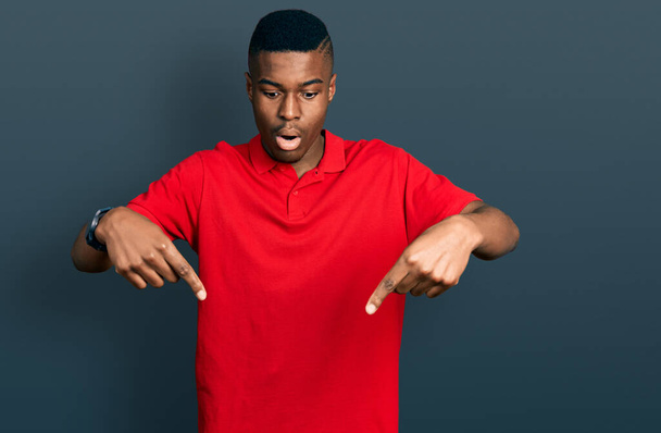Junger afrikanisch-amerikanischer Mann in lässigem rotem T-Shirt mit nach unten zeigenden Fingern, die Werbung zeigen, überraschtem Gesicht und offenem Mund  - Foto, Bild
