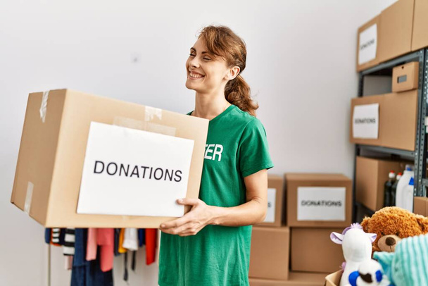 Young caucasian volunteer woman smiling happy holding donations cardboard box at charity center. - Photo, image