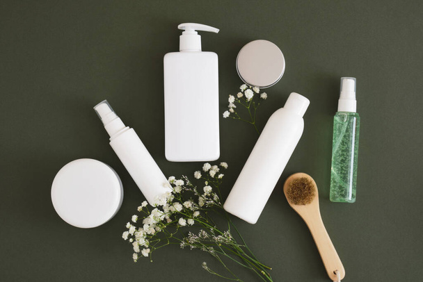 flowers, massage wooden brush, different cosmetic jars and three different blank white bottles on a dark green background backdrop Natural cosmetic packaging. mock up beauty product concept. - 写真・画像