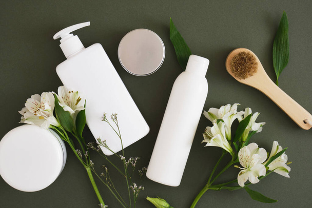 flowers, massage wooden brush, different cosmetic jars and three different blank white bottles on a dark green background backdrop Natural cosmetic packaging. mock up beauty product concept. - Photo, image