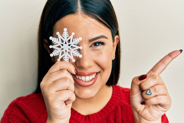 Beautiful brunette woman holding snowflake over eye smiling happy pointing with hand and finger to the side  - Photo, Image