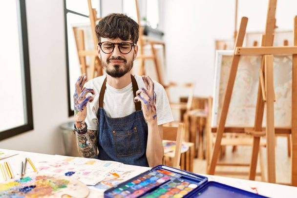 Homme hispanique avec barbe au studio d'art geste doigt croisé souriant avec espoir et les yeux fermés. chance et concept superstitieux.  - Photo, image