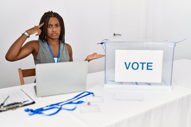 Jonge Afro-Amerikaanse vrouw die werkt bij de politieke verkiezingen zit bij stemming verward en geïrriteerd met open palm tonen kopieerruimte en wijzende vinger naar het voorhoofd. Denk er eens over na..  - Foto, afbeelding