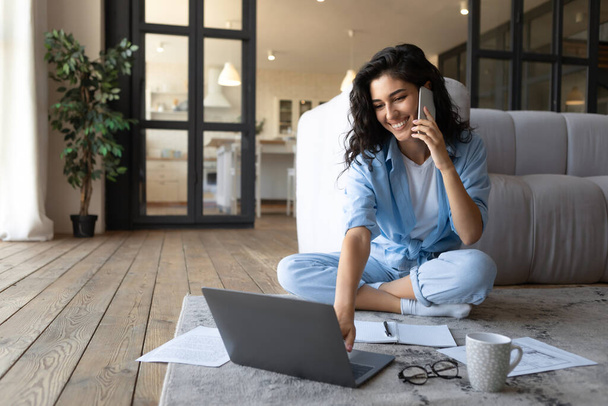 Счастливая молодая женщина, использующая ноутбук для онлайн-видеоконференции, имеющая мобильный телефон дома, копировальное пространство - Фото, изображение
