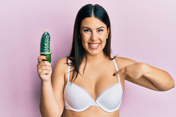 Beautiful brunette woman holding condom on cucumber for sex education pointing finger to one self smiling happy and proud  - Valokuva, kuva