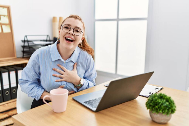 Молодая рыжая женщина, работающая в офисе с помощью ноутбука улыбается и громко смеется, потому что смешная сумасшедшая шутка с руками на теле.  - Фото, изображение