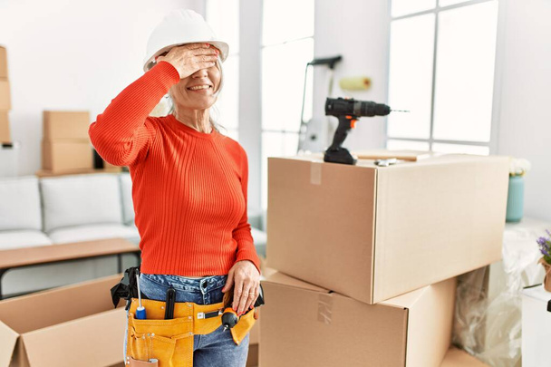 Middle age grey-haired woman wearing hardhat standing at new home smiling and laughing with hand on face covering eyes for surprise. blind concept.  - Photo, Image