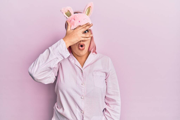 Mujer hispana con cabello rosa con máscara para dormir y pijama asomándose en shock cubriendo la cara y los ojos con la mano, mirando a través de los dedos con expresión avergonzada.  - Foto, Imagen