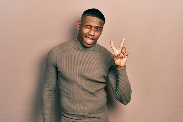 Young black man wearing casual turtleneck sweater smiling with happy face winking at the camera doing victory sign. number two.  - Photo, Image