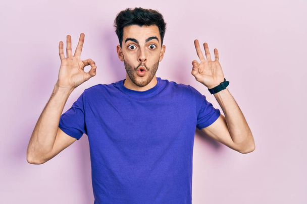 Joven hombre hispano vistiendo camiseta casual que parece sorprendido y sorprendido haciendo símbolo de aprobación aceptable con los dedos. expresión loca  - Foto, Imagen