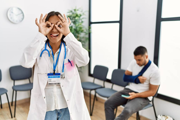 Young asian doctor woman at waiting room with a man with a broken arm doing ok gesture like binoculars sticking tongue out, eyes looking through fingers. crazy expression.  - Photo, Image