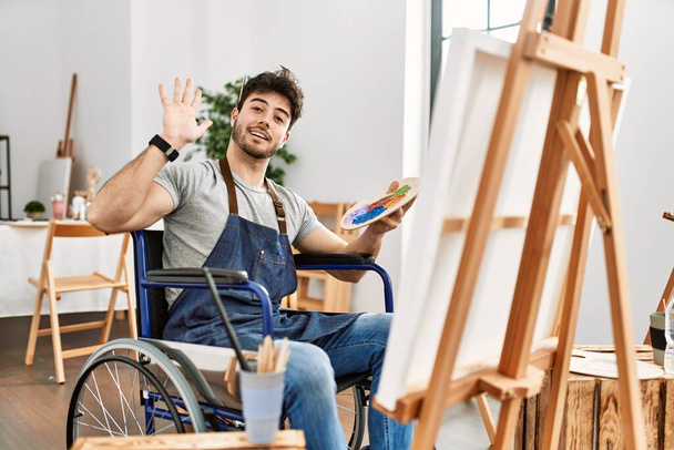Jeune homme hispanique assis sur la peinture en fauteuil roulant au studio d'art renonçant à dire bonjour heureux et souriant, geste de bienvenue convivial  - Photo, image