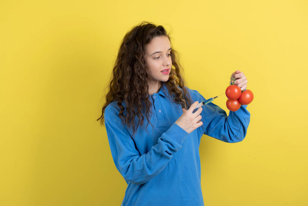 Όμορφη κοπέλα σε μπλε φούτερ έγχυση κόκκινες φρέσκες ντομάτες σε κίτρινο φόντο. Υψηλής ποιότητας φωτογραφία - Φωτογραφία, εικόνα