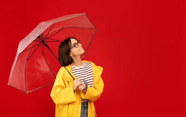 Young lady in yellow raincoat standing with umbrella and looking aside at empty space, red background - Photo, Image