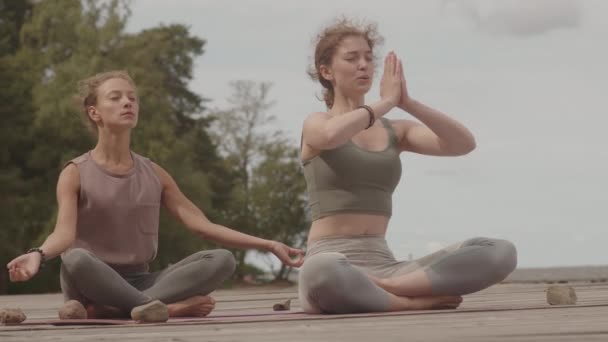 Lento disparo de dos jóvenes amigas practicando yoga juntas sentadas en un muelle de madera en un día nublado - Metraje, vídeo