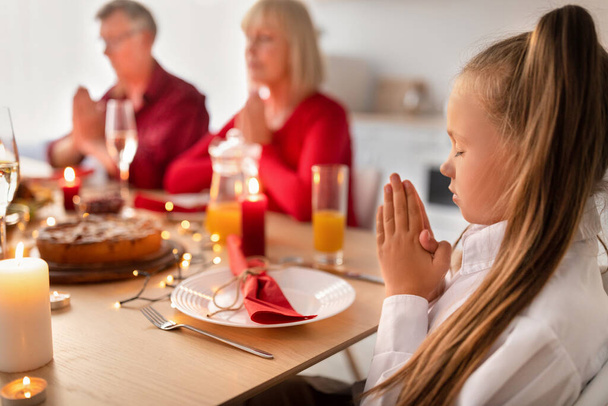 Маленька дівчинка сидить за столом, молиться з бабусею і дідусем перед сімейною вечерею, святкуючи свято зі своїми близькими вдома
 - Фото, зображення
