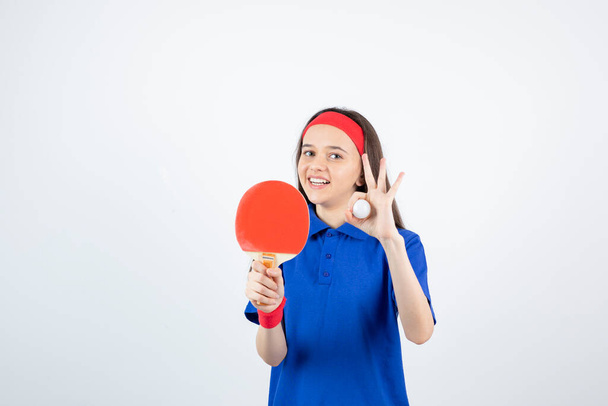 Mädchen in blauem T-Shirt, rotem Armband und Stirnband posiert mit Tennisschläger und Ball - Foto, Bild