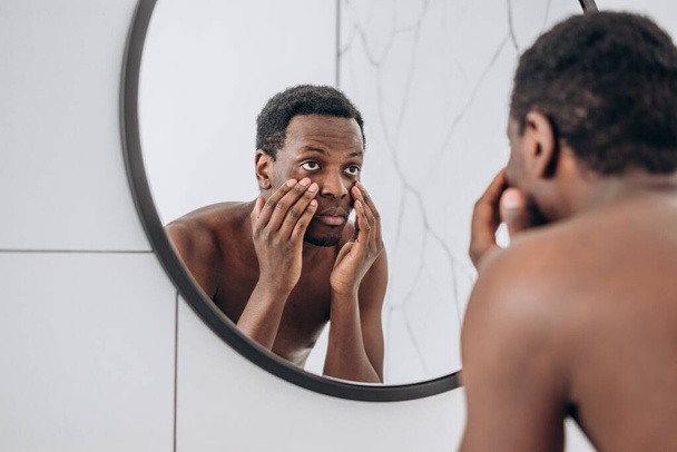 African-American man looks in mirror making face in bathroom - Foto, Imagen