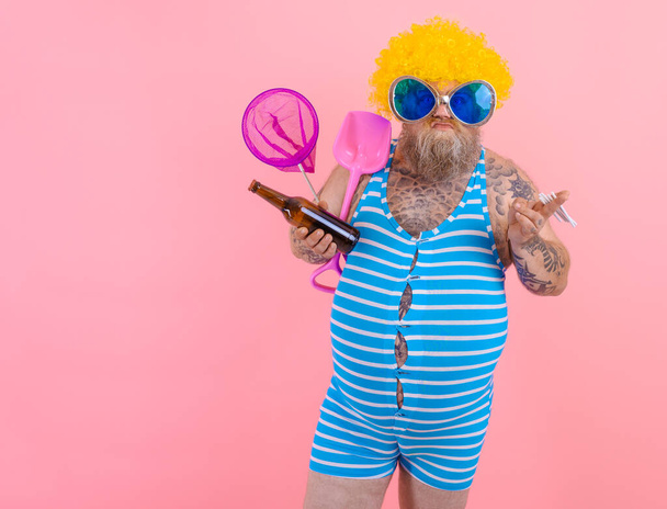 Fat man with beard and wig smokes cigarettes and drinks beer - Photo, Image