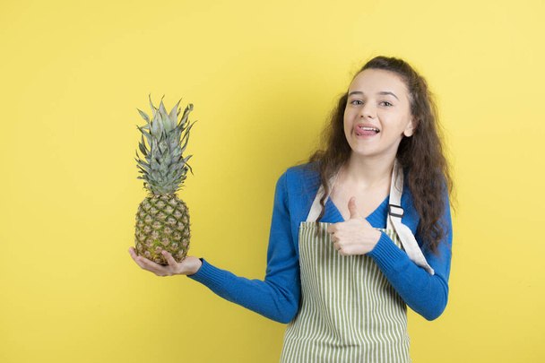 Young teen female in apron holding ripe pineapple over yellow wall. High quality photo - Photo, Image