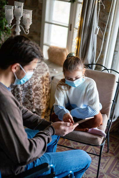 Father with disability in wheelchair using tablet at home with child while wearing masks. People education technology coronavirus concept - Photo, Image
