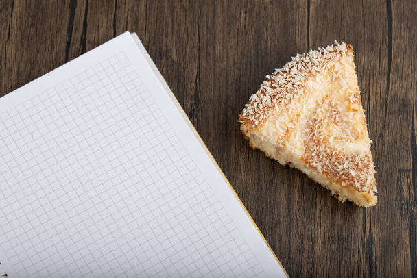 Slice of pie with coconut shavings and white notebook placed on wooden table . High quality photo - Photo, Image