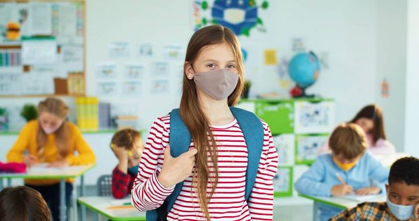 A boldog kaukázusi kislány arcmaszkot viselő portréja az osztályteremben áll, és beltéri kamerába mosolyog. A koronavírus világjárvány után tanul, többnemzetiségű általános iskolás diákok, akik a kovid időszakban tanulnak. - Fotó, kép