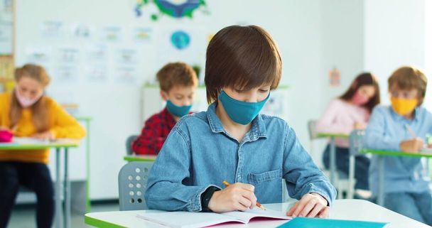 Caucasian little bright schoolboy in mask sitting at desk in classroom writing down at lesson and raising hand wants to answer question. - Valokuva, kuva