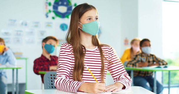 Close up portrait of Caucasian cute teen pupil girl in mask writing sitting at school classroom. Multi-ethnic children in classroom on background. Coronavirus pandemic education. Knowledge concept - Foto, Imagen