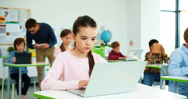 Close up of concentrated Caucasian little girl sitting at desk at school class typing on laptop Children studying in classroom using digital gadgets, computer science class modern elementary education - Φωτογραφία, εικόνα