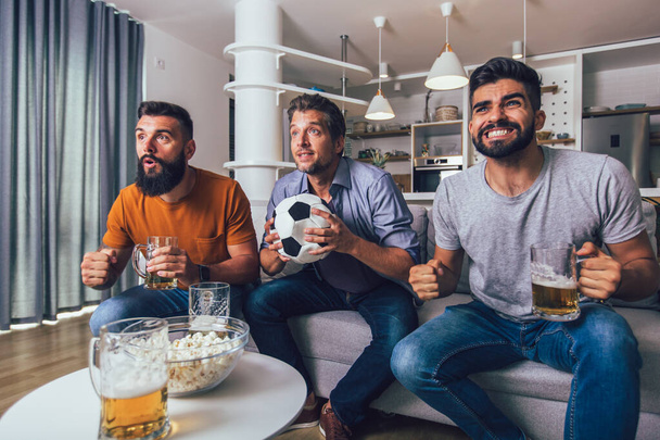 Szczęśliwy mężczyzna przyjaciele lub kibice piłki nożnej oglądając piłkę nożną w telewizji i świętując zwycięstwo w domu. - Zdjęcie, obraz