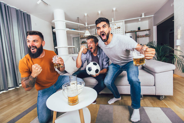 Ευτυχισμένοι άνδρες φίλοι ή οπαδοί του ποδοσφαίρου βλέποντας ποδόσφαιρο στην τηλεόραση και γιορτάζοντας τη νίκη στο σπίτι. - Φωτογραφία, εικόνα