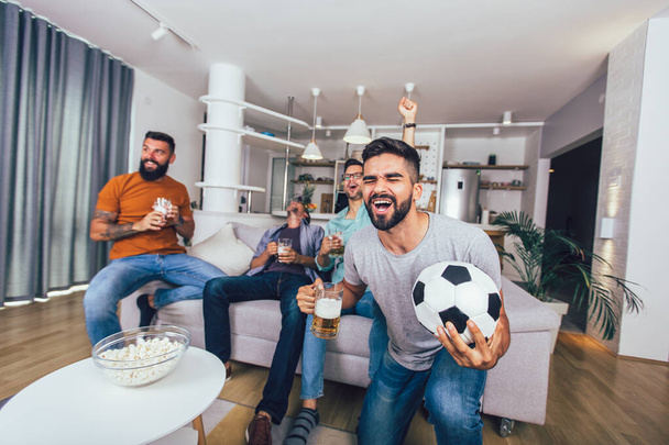 家で一緒にテレビでスポーツを見ている男性は陽気に叫んでいる。ソファに座ってサッカーの試合を見ている友人のグループ. - 写真・画像
