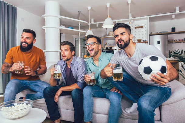 家で一緒にテレビでスポーツを見ている男性は陽気に叫んでいる。ソファに座ってサッカーの試合を見ている友人のグループ. - 写真・画像