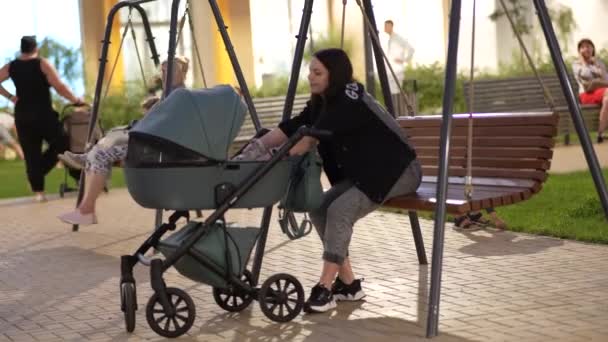 Mom with a stroller is sitting on a swing in the courtyard of a residential building - Footage, Video