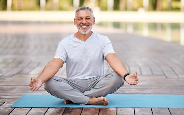 Spokojny, szczęśliwy starszy mężczyzna siedzący w lotosowej pozie na macie podczas porannej medytacji w parku, trzymający się za ręce w geście mudry i uśmiechający się do kamery. Zdrowie psychiczne i koncepcja jogi - Zdjęcie, obraz