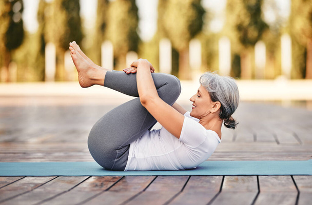 Вид сбоку зрелой женщины в спортивной одежде, лежащей на коврике, обнимающей колени к груди после занятий йогой на открытом воздухе в парке, здоровая активная пожилая женщина, делающая утром упражнения на растяжку на свежем воздухе - Фото, изображение