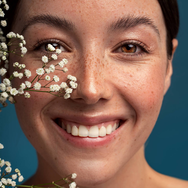 Beauty natural smiling woman with freckles, clean and healthy glowing skin. Concept of sunscreen lotions, skincare cosmetics and spa. Girl looking at the camera. Happy emotions. Small white flowers on her face. Studio shot. - Fotografie, Obrázek