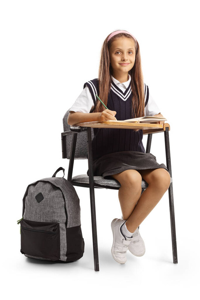Girl in a school uniform sitting in a school chair with a pen and a notebook and looking at camera isolated on white background - Photo, image