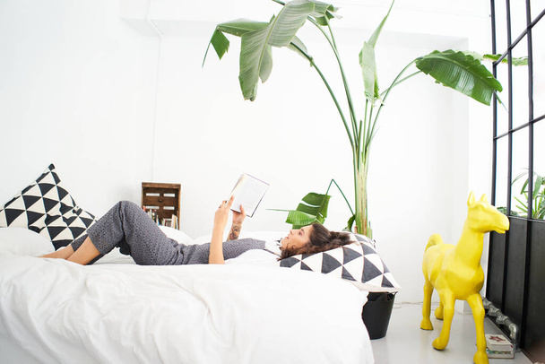 Jeune latina lit un livre dans la chambre couchée sur le lit profitant d'un nouveau jour, se sentant reposée et heureuse. - Photo, image