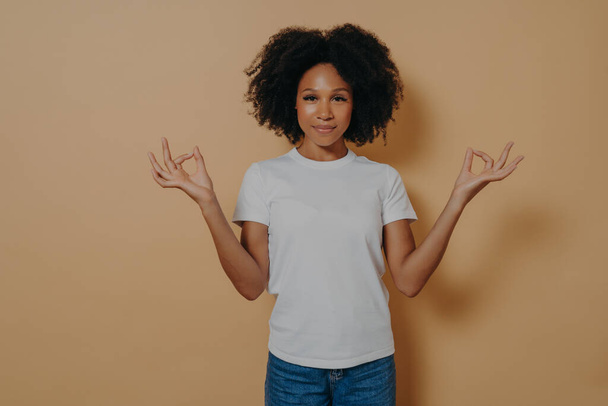 Ragazza afro-americana pacifica che tiene le mani nel gesto mudra, isolata sulla parete beige - Foto, immagini