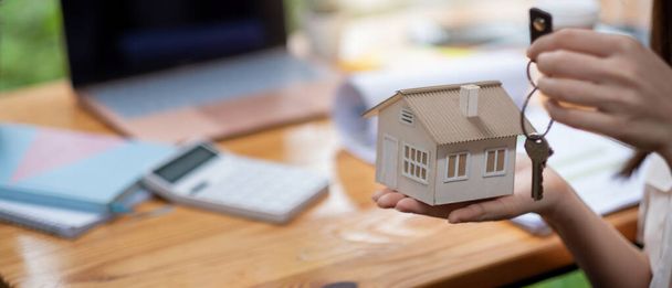 hipoteca, bienes raíces y concepto de propiedad - primer plano de las manos que sostienen el modelo de casa y las llaves del hogar con fondo de espacio de trabajo financiero. - Foto, imagen