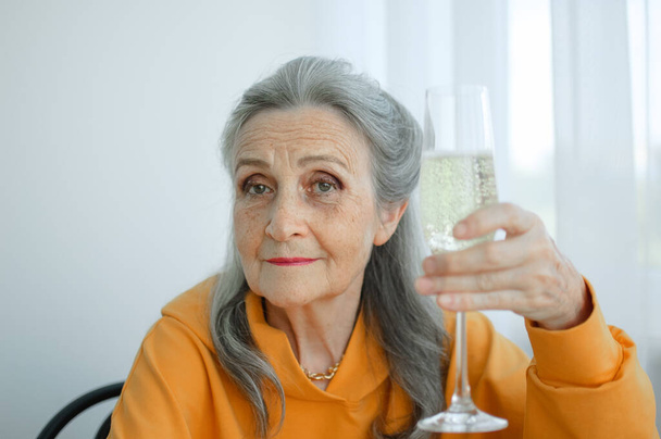 Beautiful old grandmother with grey hair and face with wrinkles sitting at the table at home on window background with glass of champagne, mothers day, happy retirement - Foto, Imagen