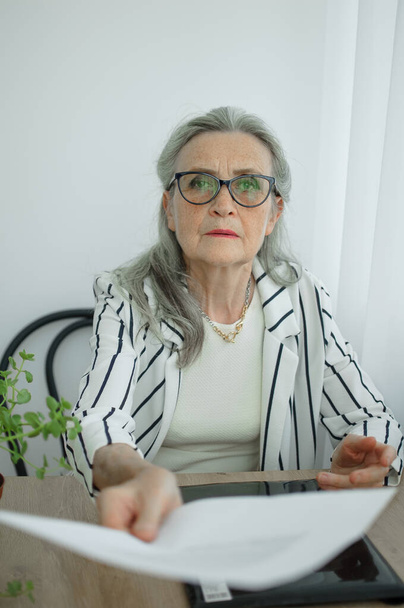 Senior grey haired businesswoman in striped jacket with eyeglasses is working with documents in here office sitting at the desk. Happy retirement, employment and labor - Foto, Bild