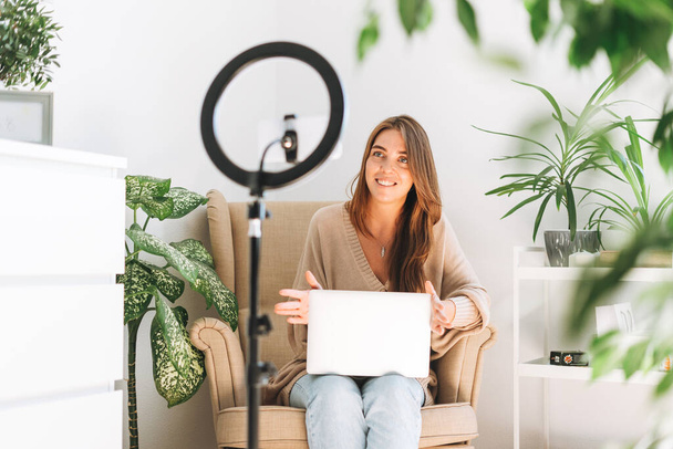Nuori kaunis nainen bloggaaja tallentaa videon matkapuhelimeen käyttämällä rengaslamppua, joka istuu tuolilla kannettavalla tietokoneella huoneessa, jossa on vihreitä kasveja kotona - Valokuva, kuva