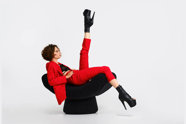 Flexible woman in red pantsui and pole dance boots sits in a soft armchair and lift one leg up - Foto, afbeelding