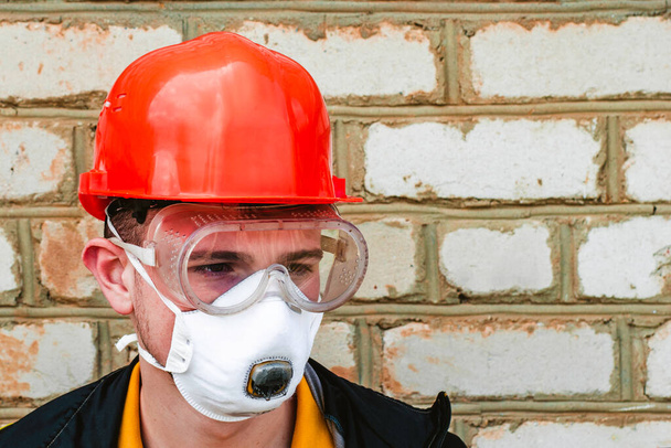 Парень строитель в защитном строительном шлеме респираторе и защитных очках на фоне кирпичной стены копирайта. Высокое качество фото - Фото, изображение