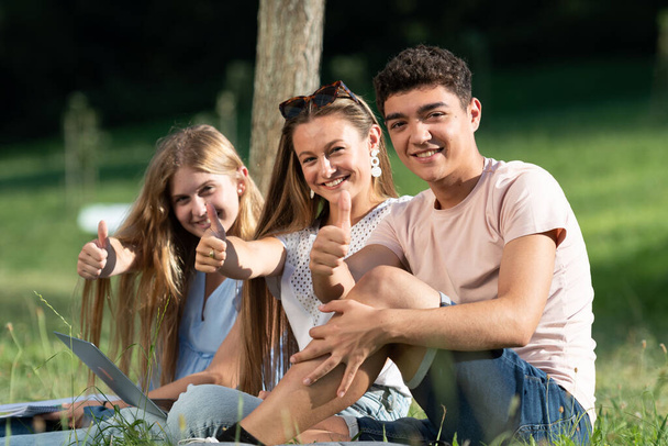 Studenti felici di successo gesticolando pollici in su seduti all'aperto sul terreno di un parco. - Foto, immagini